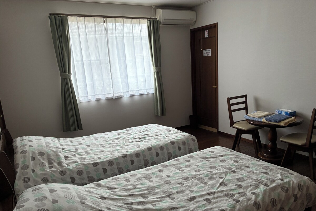 Ein Zimmer mit zwei Betten, einem Tisch mit Stühlen, einem Fenster mit Vorhängen und einer Tür mit einem Schild.