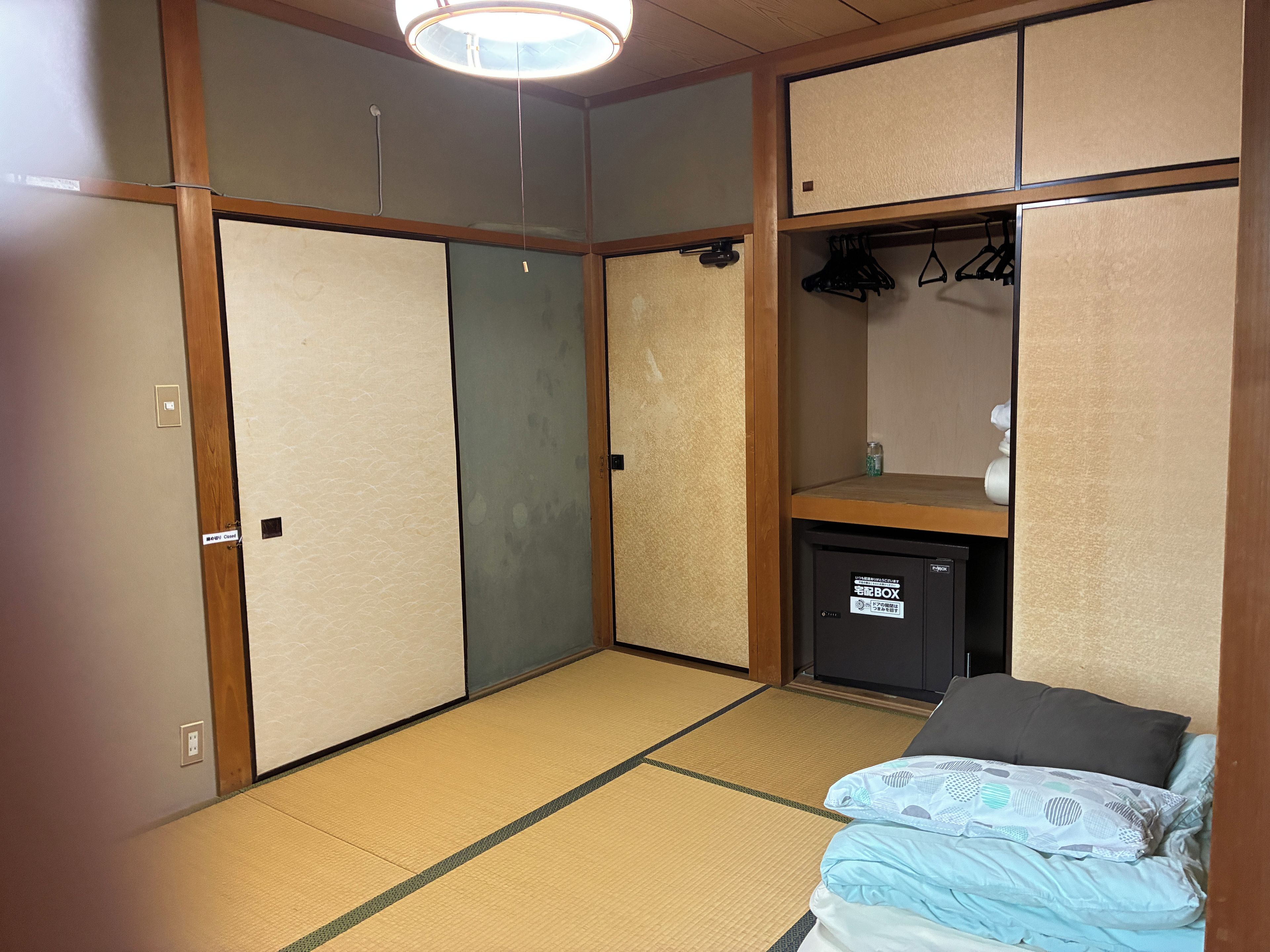 A room with tatami flooring, a bed with a patterned blanket, a small cabinet, and a ceiling light fixture.