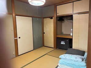 A room with tatami flooring, a bed with a patterned blanket, a small cabinet, and a ceiling light fixture.