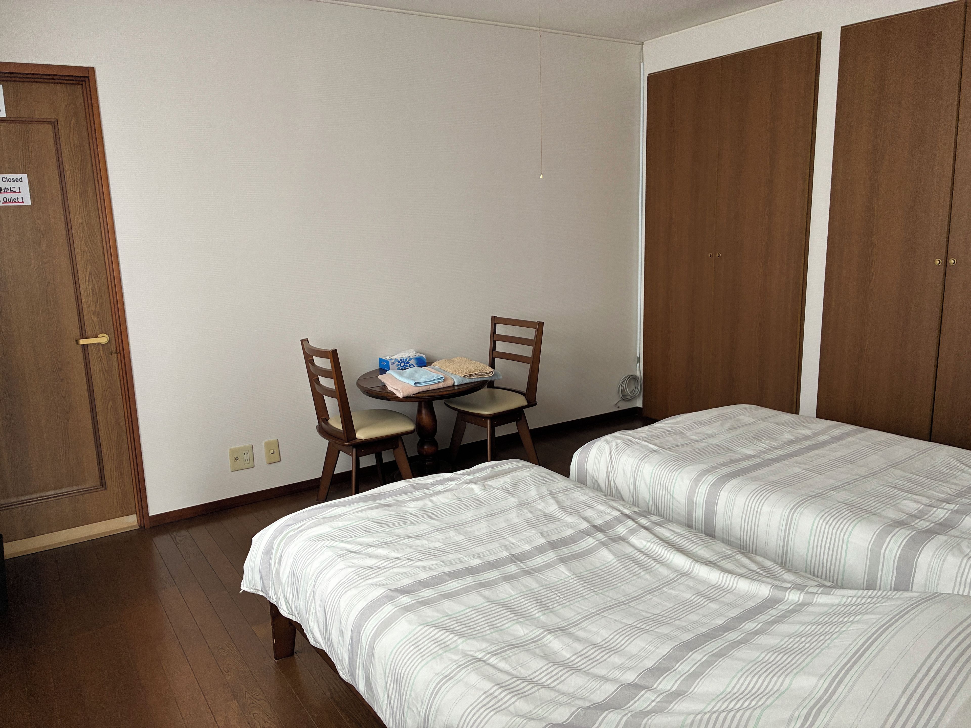A hotel room with two beds, a small table with a chair, and a wardrobe.