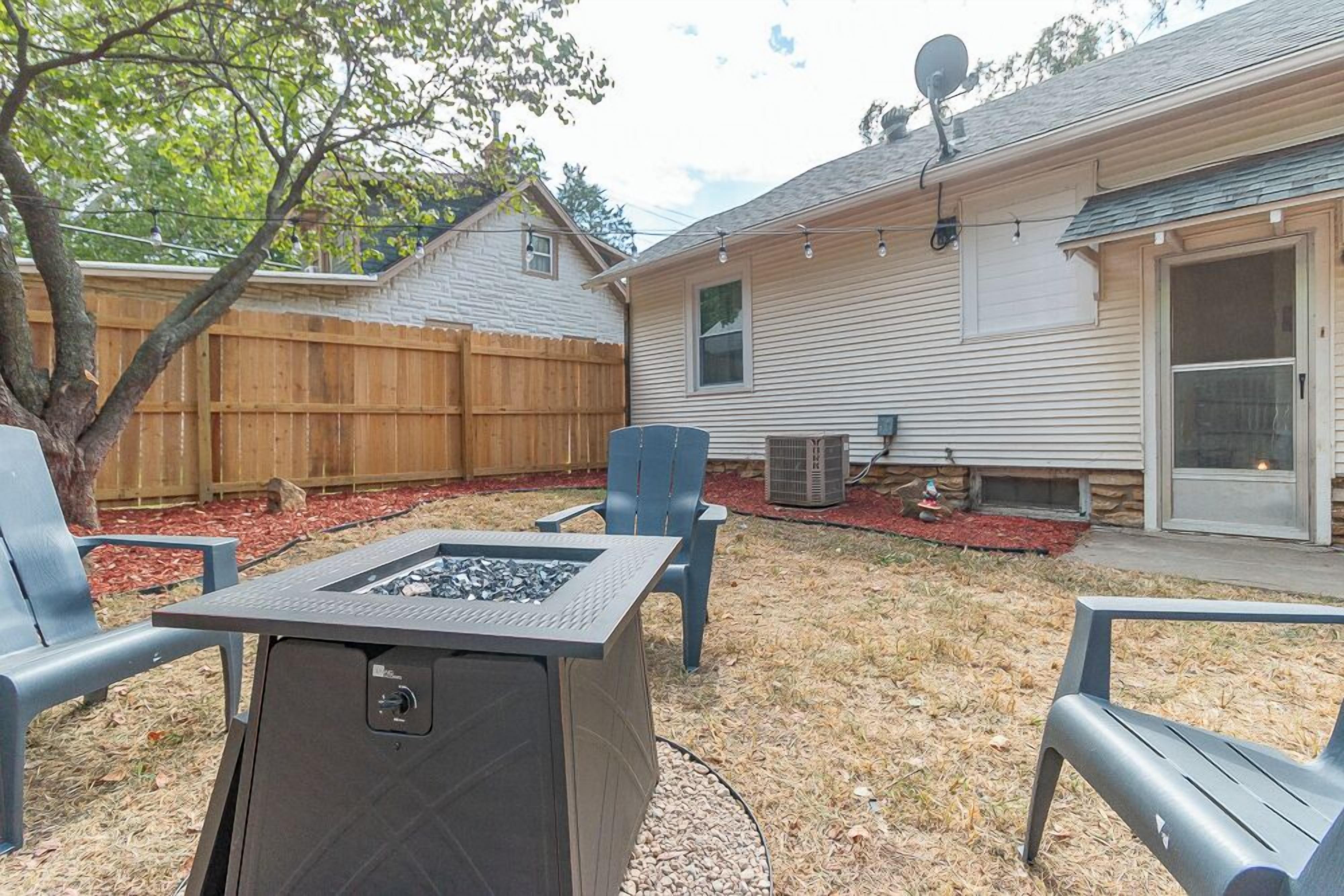 A backyard with a fire pit, outdoor furniture, a wooden fence, and a house with a satellite dish.