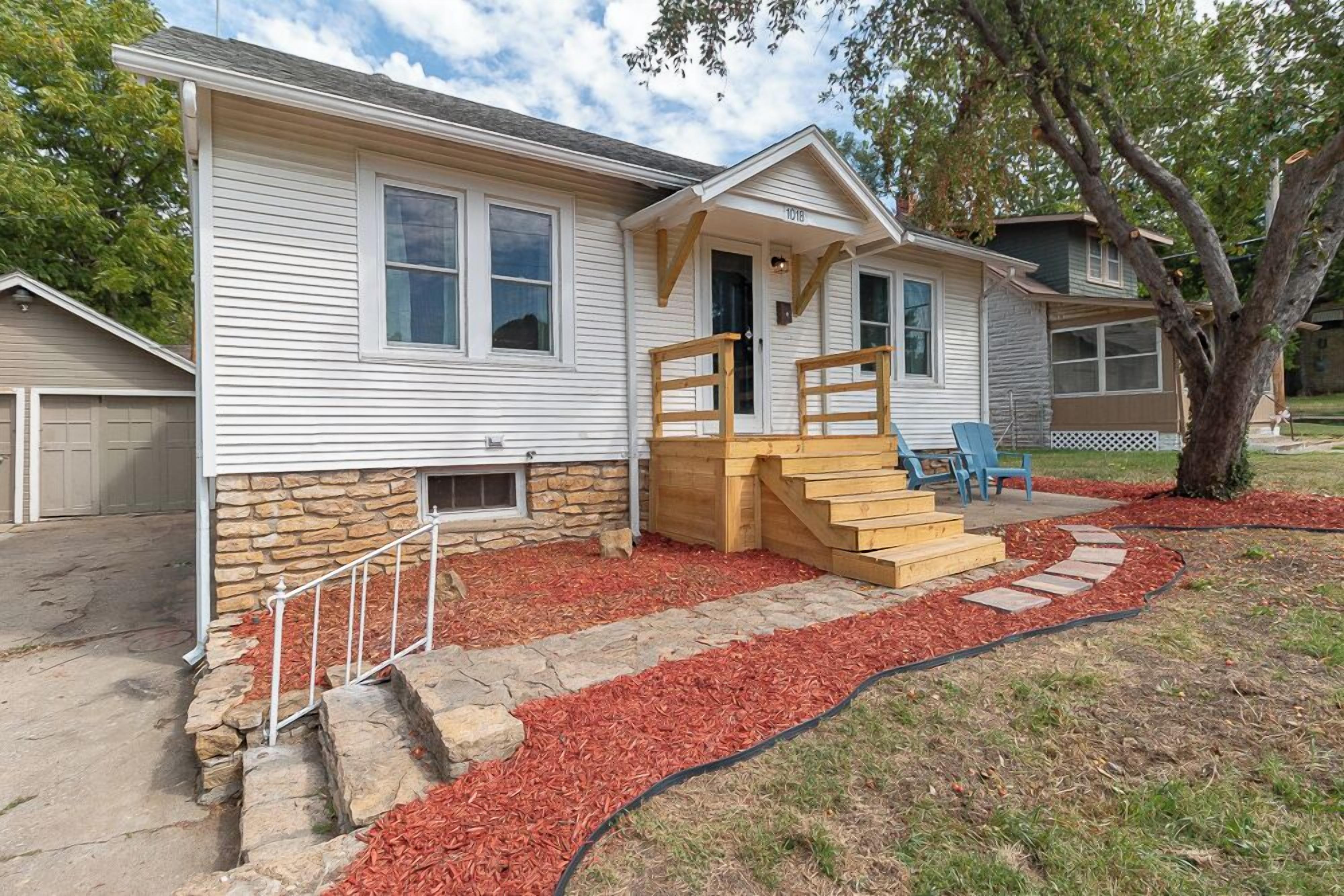 A single-story house with a stone foundation, a front porch with a wooden staircase, and a red mulch pathway leading to the entrance.
