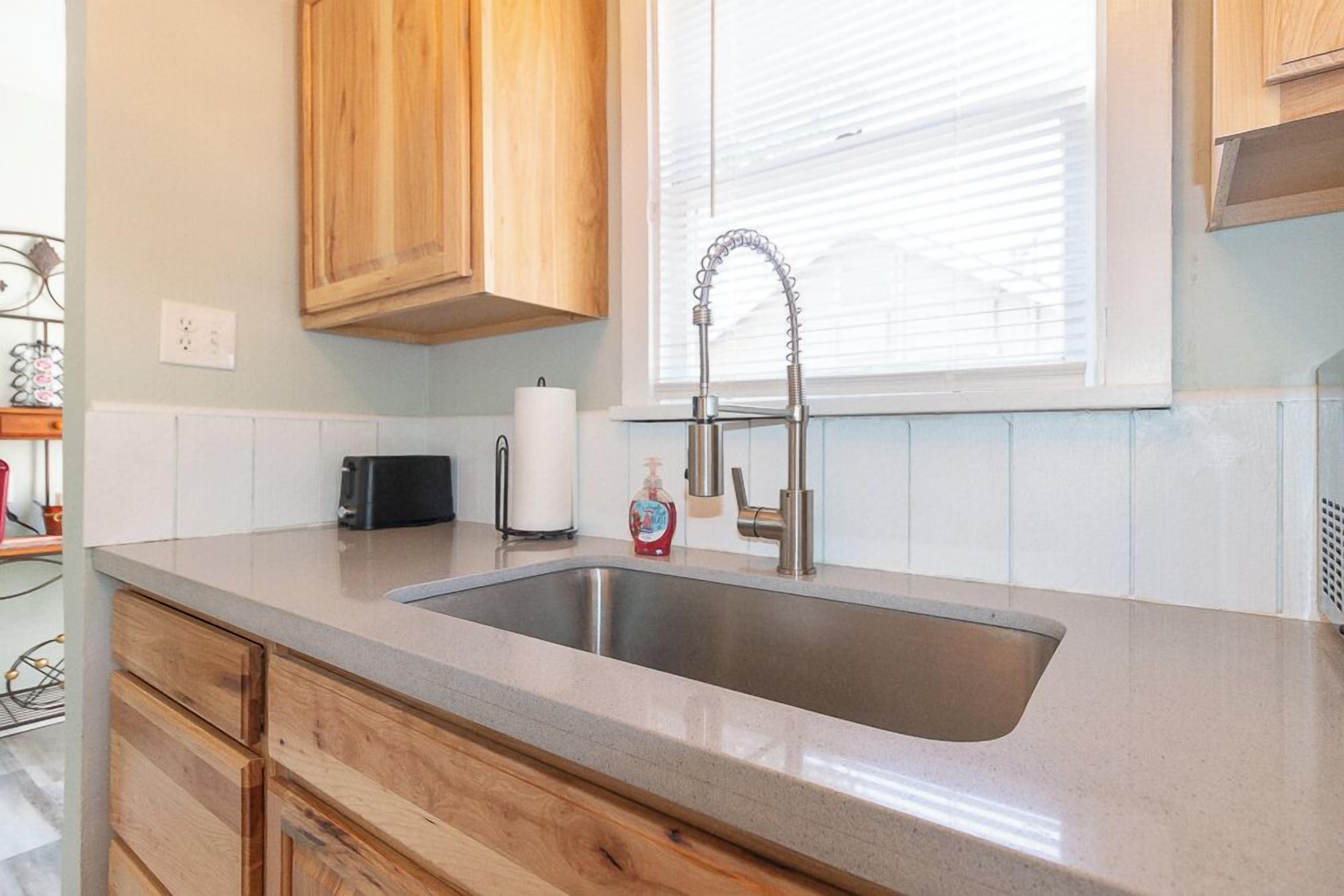 A kitchen with a stainless steel sink, wooden cabinets, a microwave, and a window with blinds.