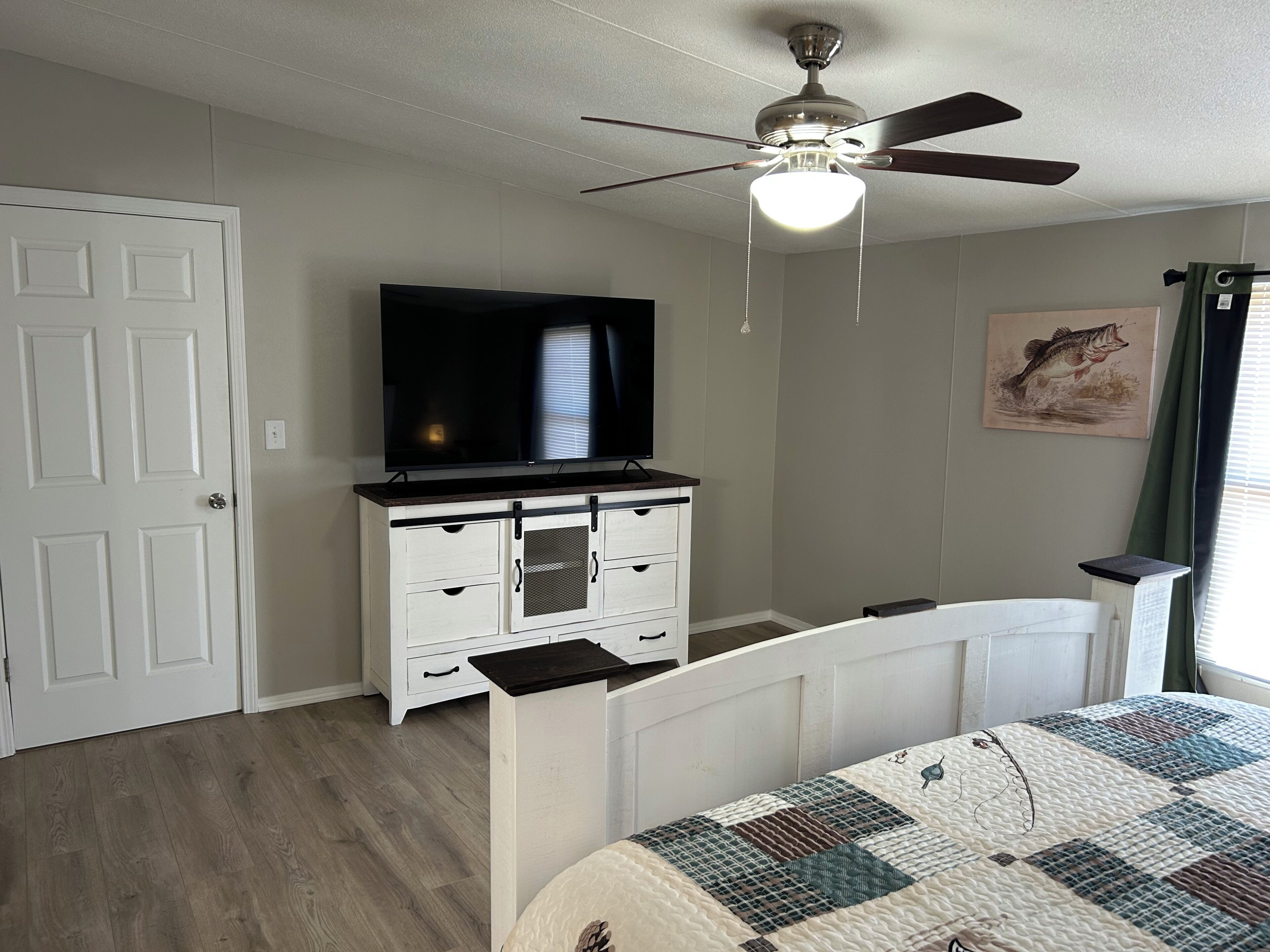 A bedroom with a bed, a television on a dresser, a ceiling fan, and a painting on the wall.