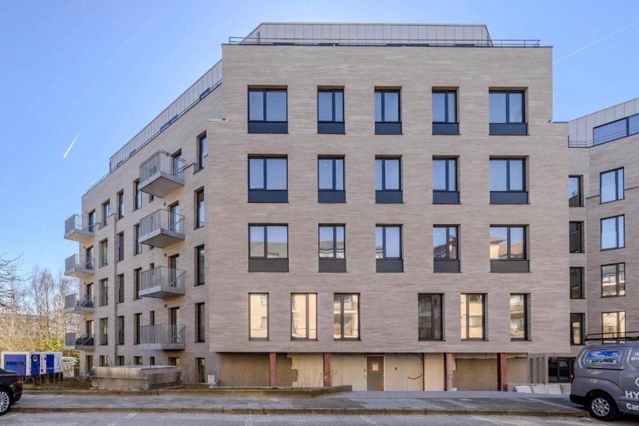 Ein mehrgeschossiges Gebäude mit flacher Fassade, Balkonen und einem Parkplatz mit einem Fahrzeug.