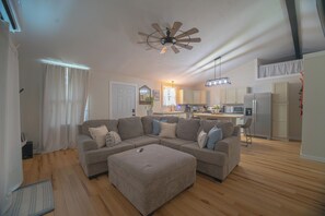 Ein modernes Wohnzimmer mit einem Ecksofa, einem Deckenventilator und einer Küche im Hintergrund.