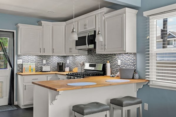 A modern kitchen with white cabinets, a wooden countertop, a gas stove, a microwave, and a toaster. There are two stools and a window with blinds. - Beach Cottage Gem - 2Blocks2 Beach, Parking, Cozy (Huntington Beach)
