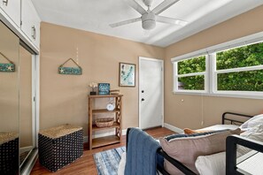 A bedroom with a bed, a window with a view of greenery, a ceiling fan, and a shelf with decorative items.