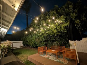 An outdoor patio area with a wooden table and chairs, surrounded by a white fence and greenery.