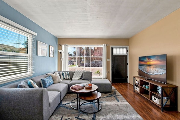 A living room with a sectional sofa, a coffee table, a TV on a wooden stand, and a view through a glass door.