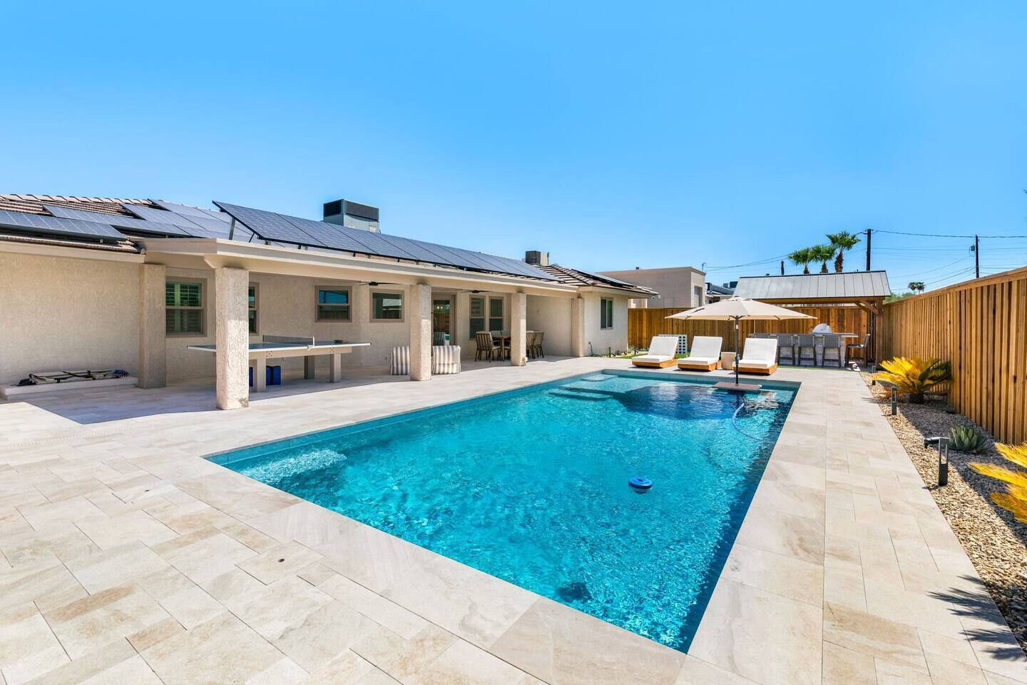 Une piscine rectangulaire, bordée d’une terrasse carrelée, entourée d’une clôture en bois et d’un bâtiment doté d’un toit en pente.
