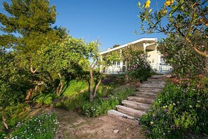 A house with a wooden staircase leading up to a white porch, surrounded by lush greenery and flowering plants.