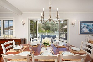 A dining room with a large wooden table set for six, white chairs, a chandelier, and a view through large windows to an outdoor deck.