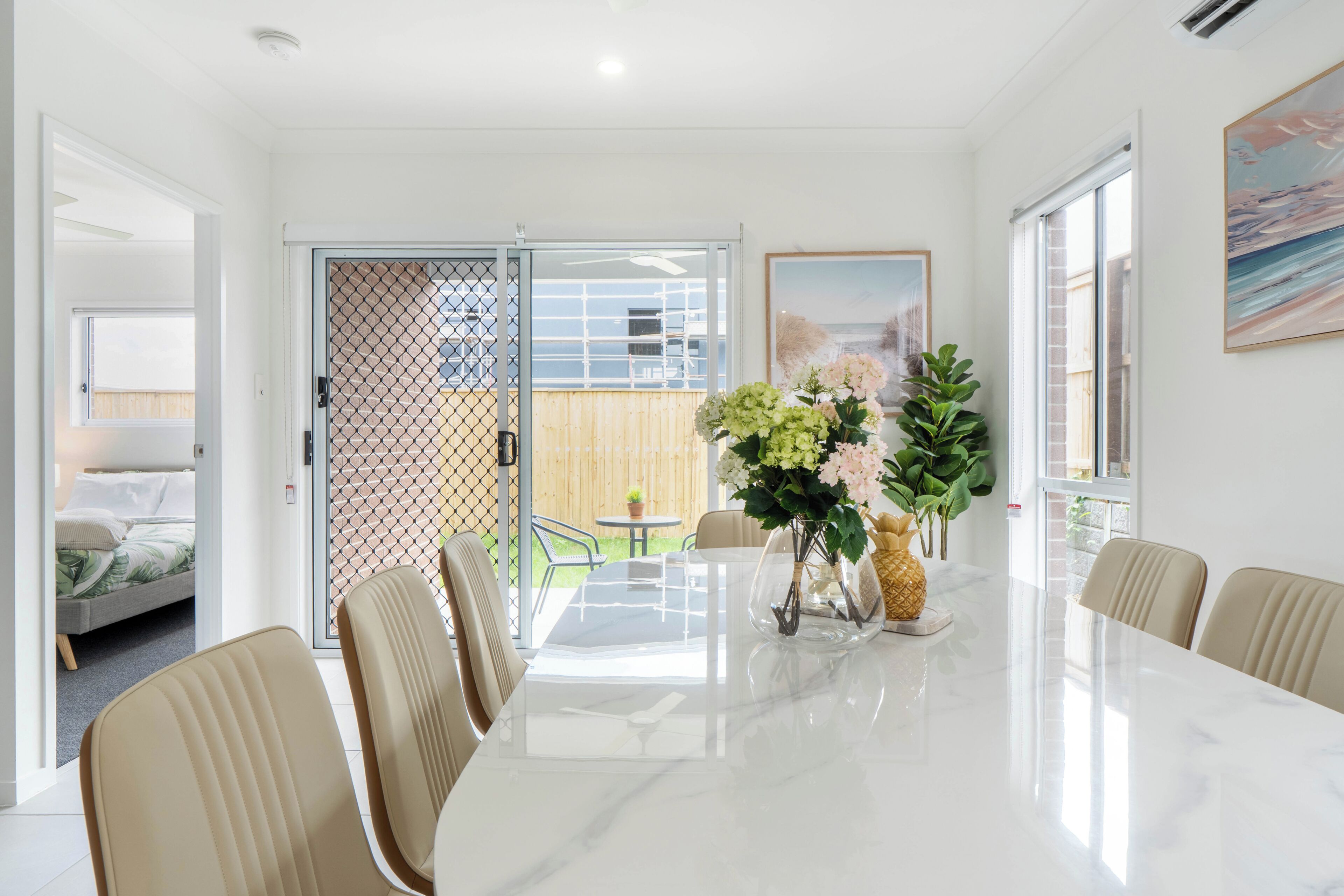 A modern dining area with a marble table, beige chairs, and a view of the backyard through a glass door.