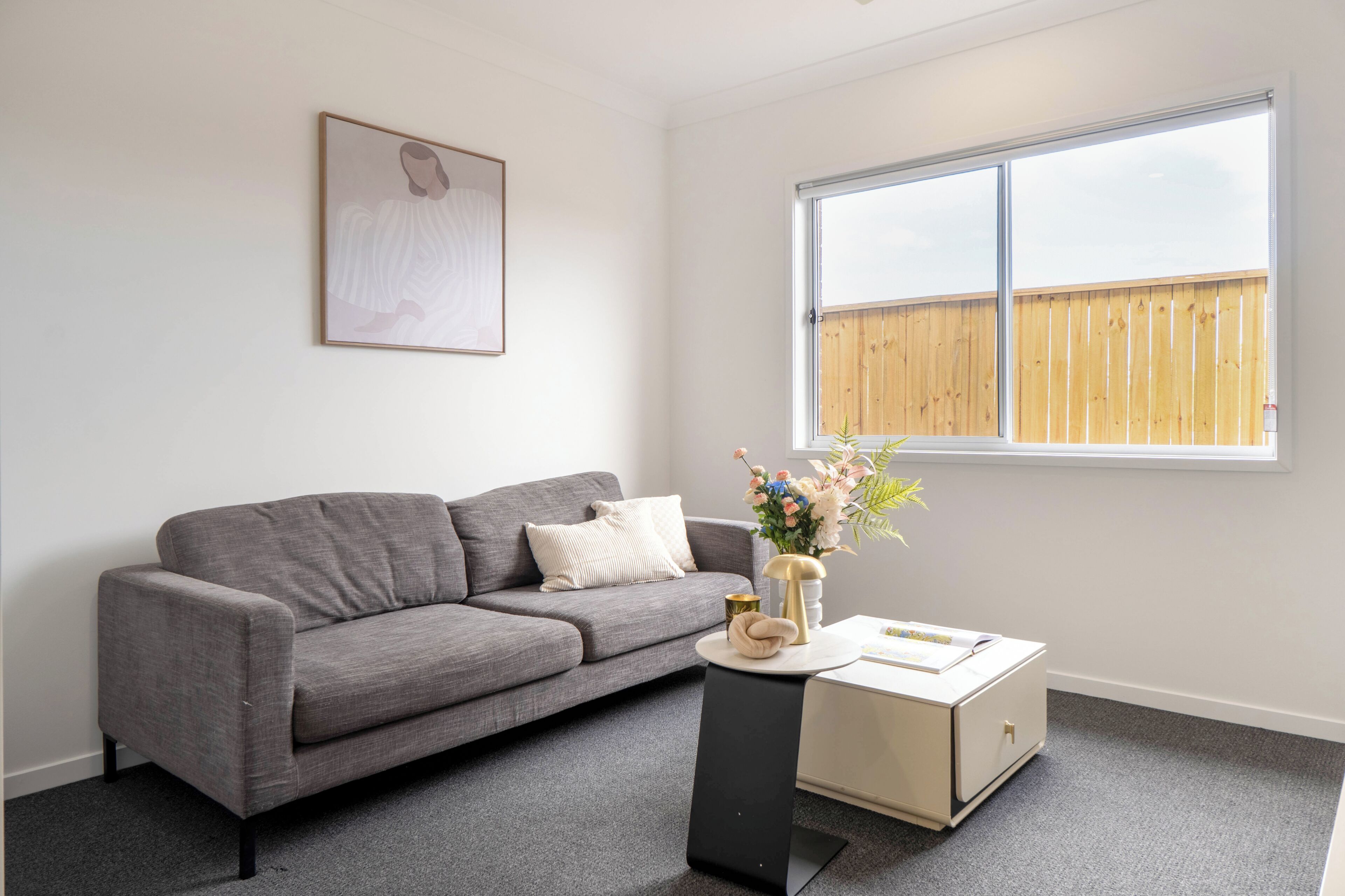 A living room with a grey sofa, a small side table, a vase with flowers, and a window with a wooden fence outside.