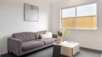 A living room with a grey sofa, a small side table, a vase with flowers, and a window with a wooden fence outside.