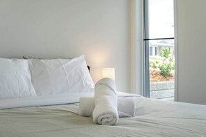 Una cama bien hecha con sábanas blancas y dos toallas enrolladas, una lámpara de noche y una ventana con vistas a zonas verdes.