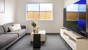 Un salon moderne avec un canapé gris, une table basse agrémentée d’objets décoratifs, un grand téléviseur à écran plat et une fenêtre donnant sur une clôture en bois.