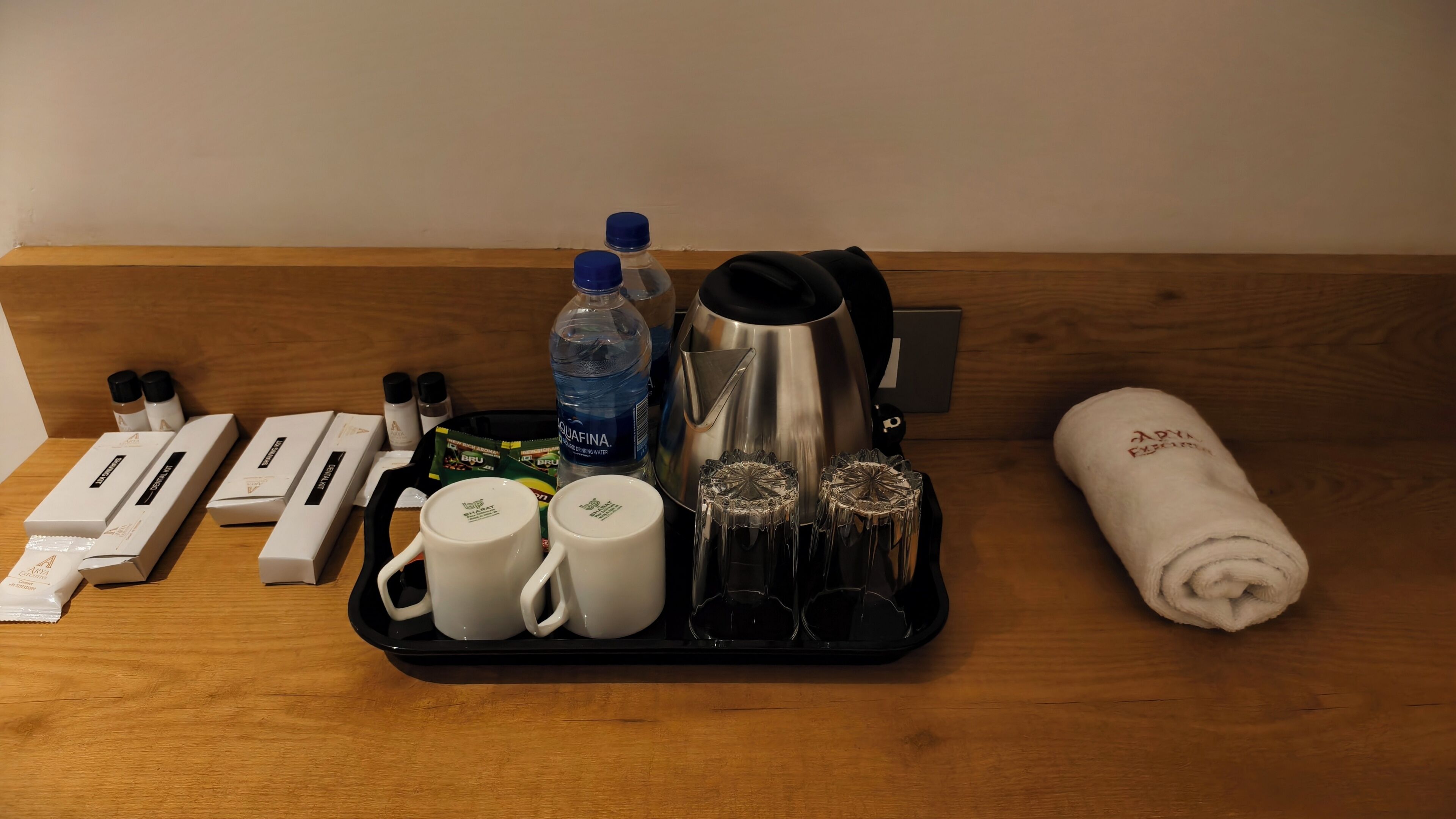 A tray with toiletries, a water bottle, a kettle, and a rolled towel on a wooden surface.