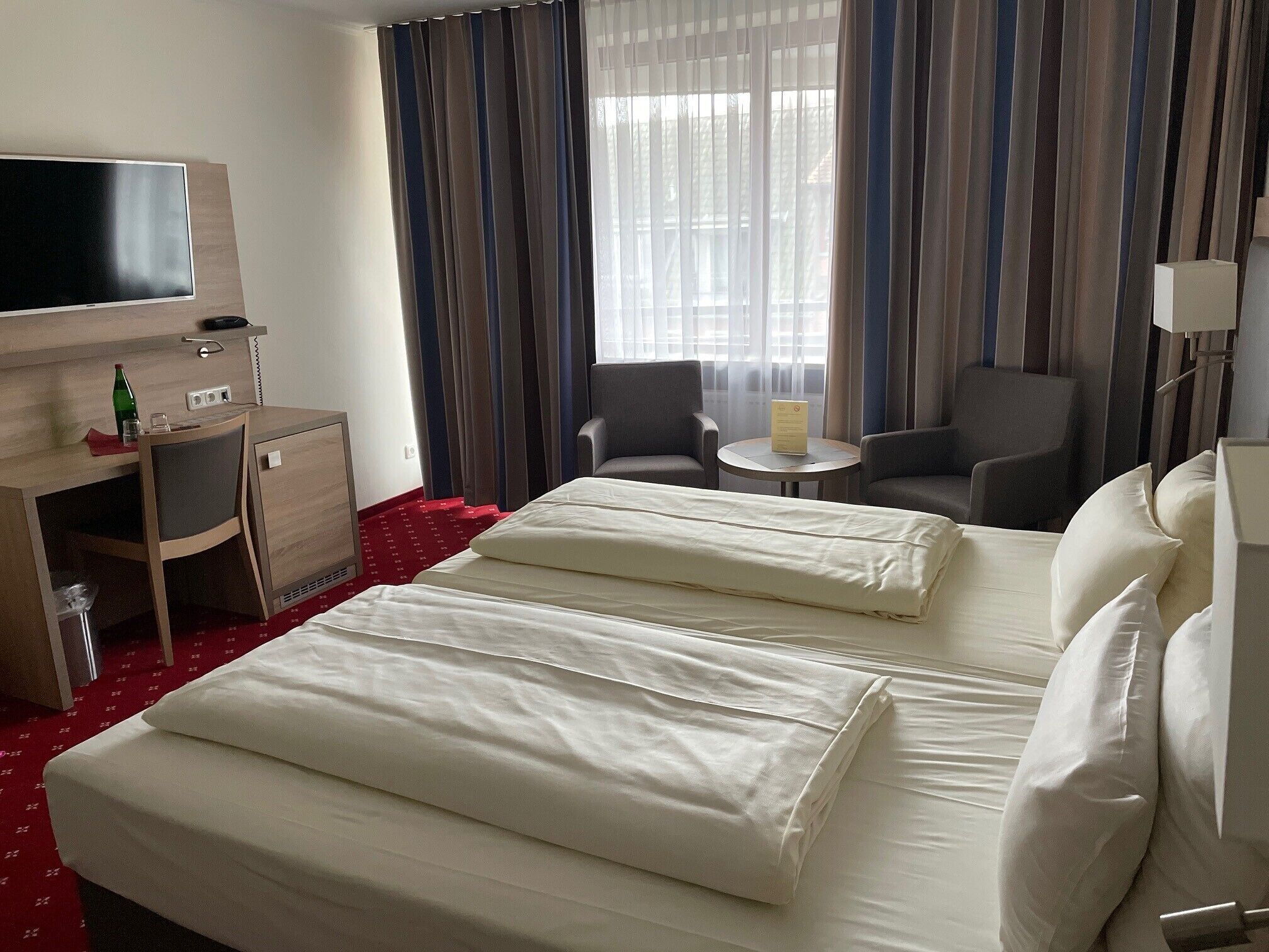 Una habitación de hotel con cama, escritorio, silla, televisión y ventana con cortinas.