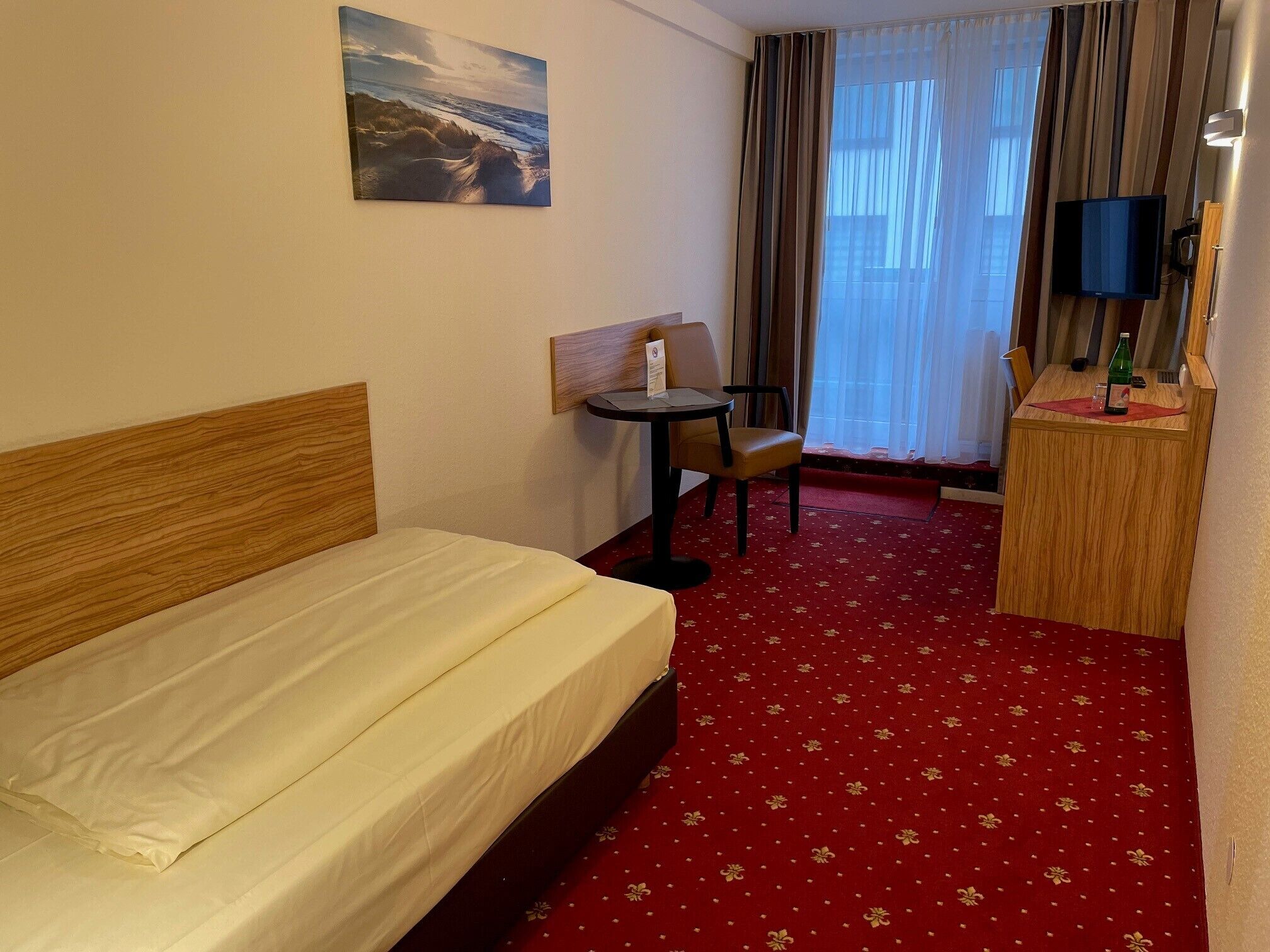 A hotel room with a bed, a desk, a chair, a television, and a painting on the wall.