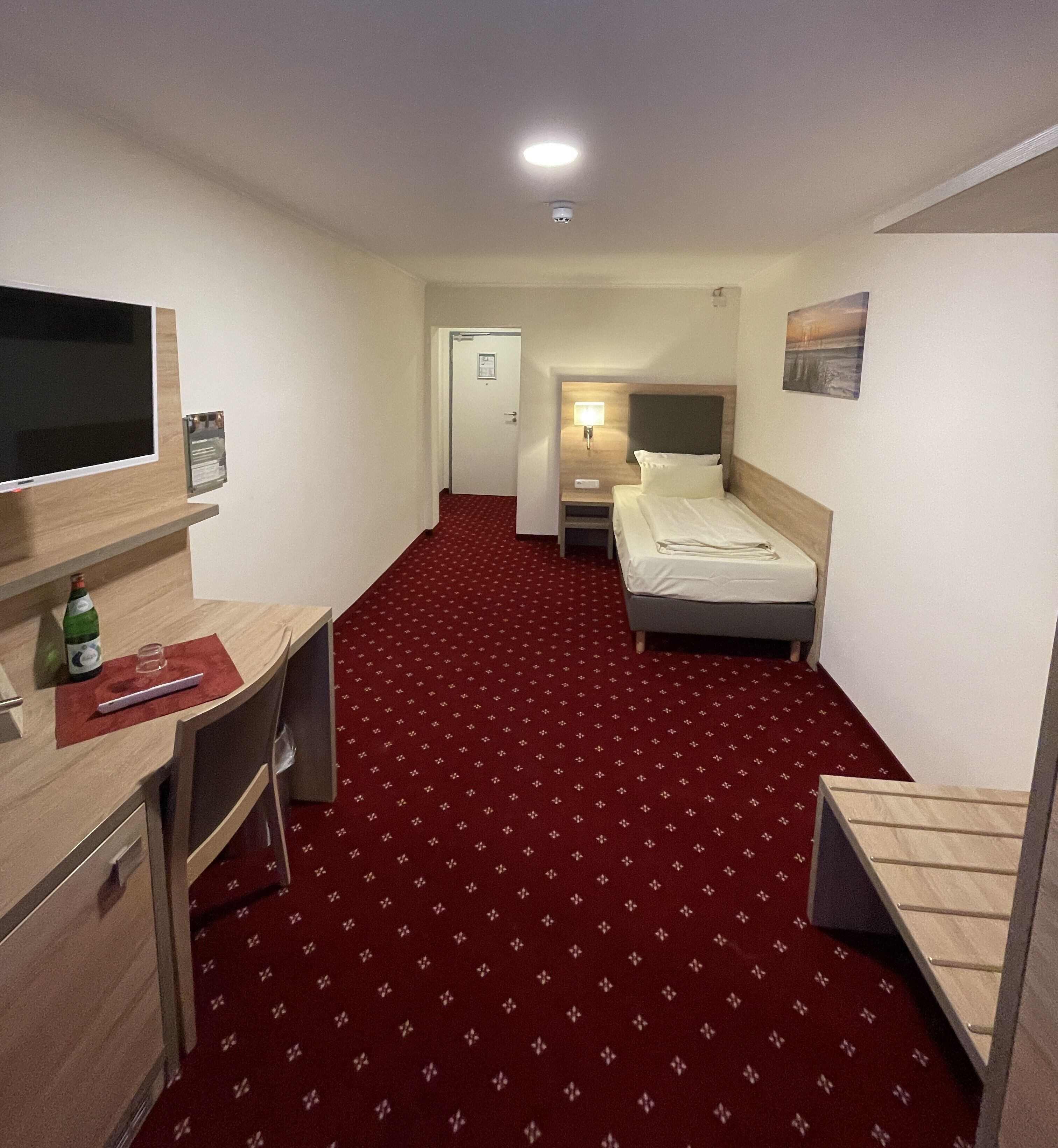 A hotel room with a bed, a desk, a television, and a red carpeted floor.
