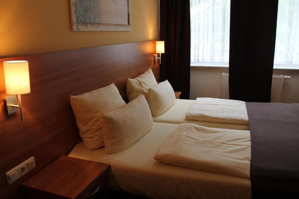 Quarto de hotel com uma cama de casal, duas luminárias de cabeceira, mesa de cabeceira e uma janela com cortinas.