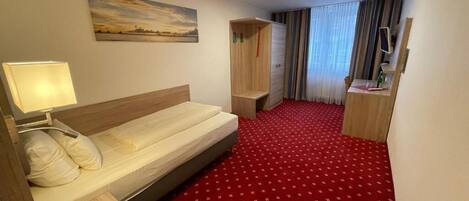 A hotel room with a bed, a desk, a mirror, a red carpet, and a painting on the wall.