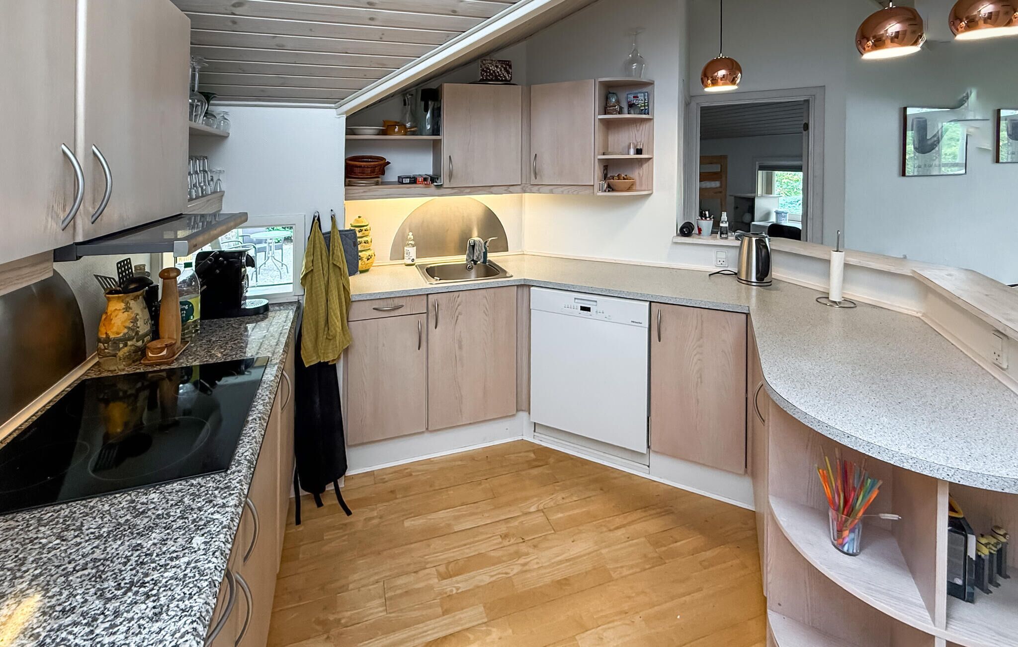 A modern kitchen with a dishwasher, granite countertops, wooden cabinets, and a view into the dining area.