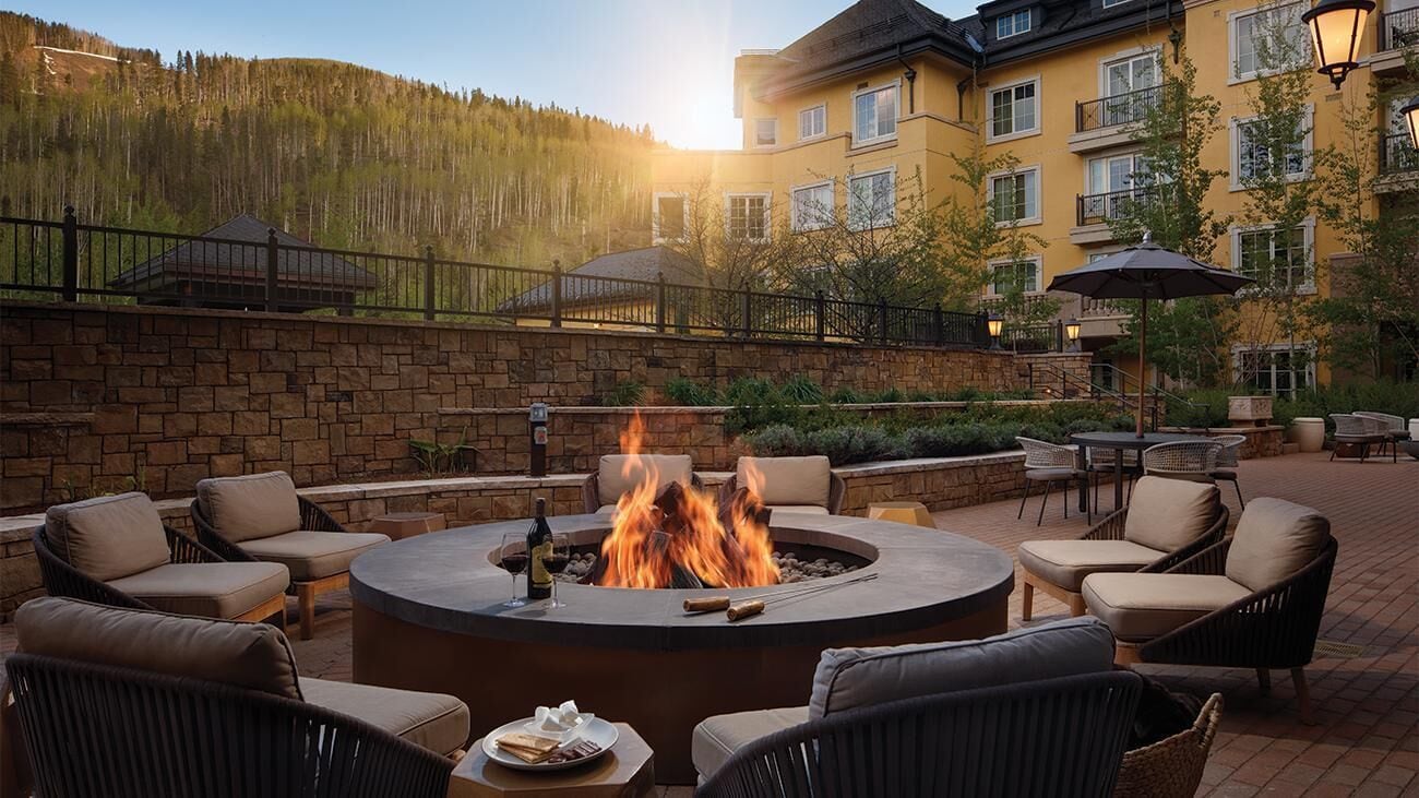 A circular fire pit surrounded by wicker chairs and a table, with a building in the background.