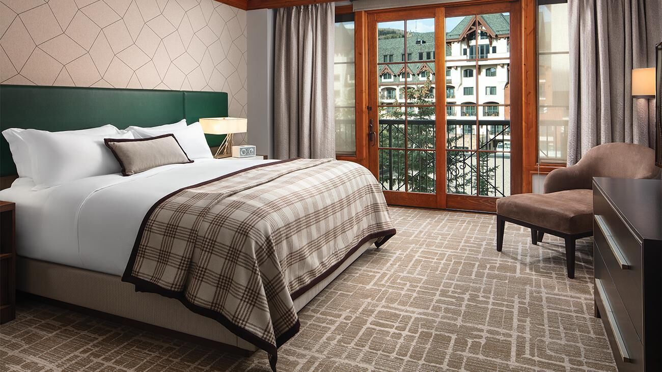 Habitación de hotel con una cama grande, una mesita de noche, una silla, un aparador y un balcón con vistas a edificios.