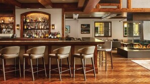 A bar area with a wooden bar counter, multiple stools, and a variety of bottles. There are chairs and a fireplace in the background. - The Ritz-Carlton Club -- 2 bedroom -- Vail, Colorado (Vail)
