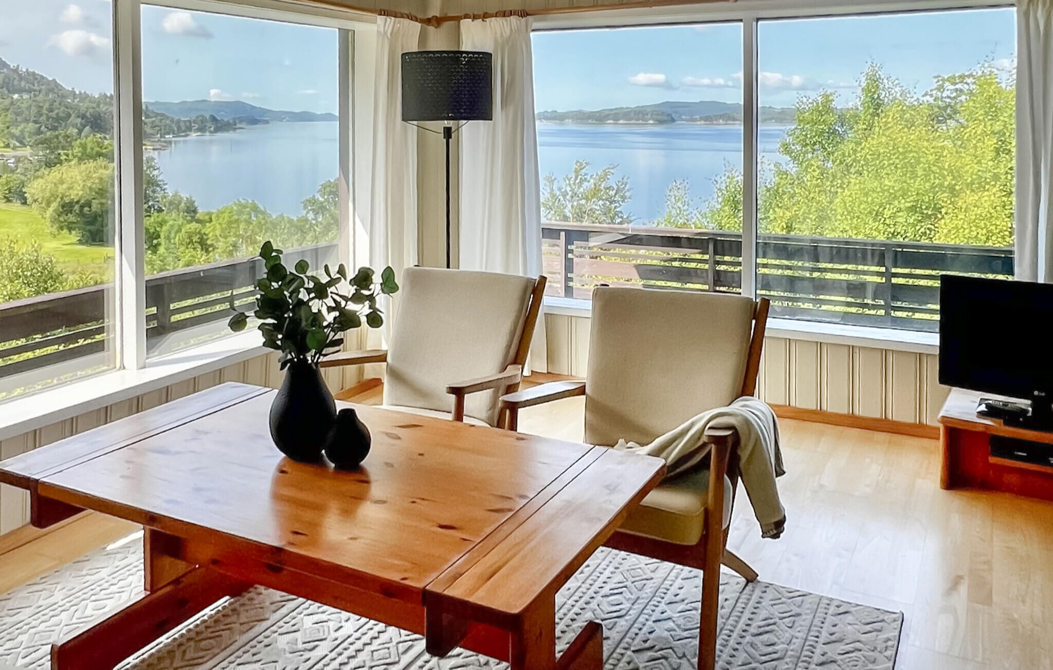 Una habitación con una mesa de madera, dos sillas, un televisor y un ventanal con vistas a un lago y colinas.