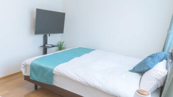 Una cama individual con una colcha verde azulada, ropa de cama blanca y una almohada azul. Un televisor de pantalla plana está montado en la pared sobre la cama.