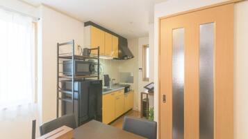 Cocina pequeña con microondas, estufa y armarios amarillos. Se observa una mesa de comedor con sillas y una puerta de madera con paneles de vidrio esmerilado.