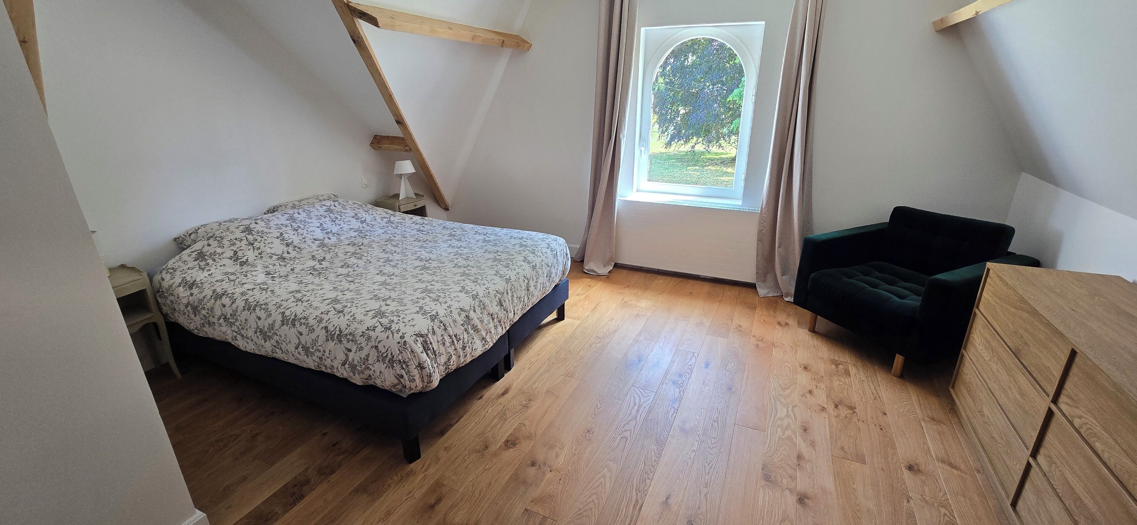 Ein Schlafzimmer mit einem Bett, einem Sessel, einer Holzkommode und einem Fenster mit Vorhängen.