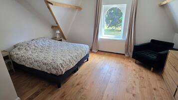 Ein Schlafzimmer mit einem Bett, einem Sessel, einer Holzkommode und einem Fenster mit Vorhängen.