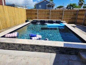 Una piscina rectangular con borde de piedra, cerca de madera y flotador inflable azul.