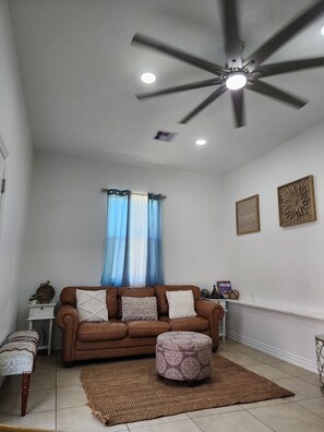 Uma sala de estar com um sofá de couro marrom, um banquinho redondo, uma pequena mesa branca e um ventilador de teto.