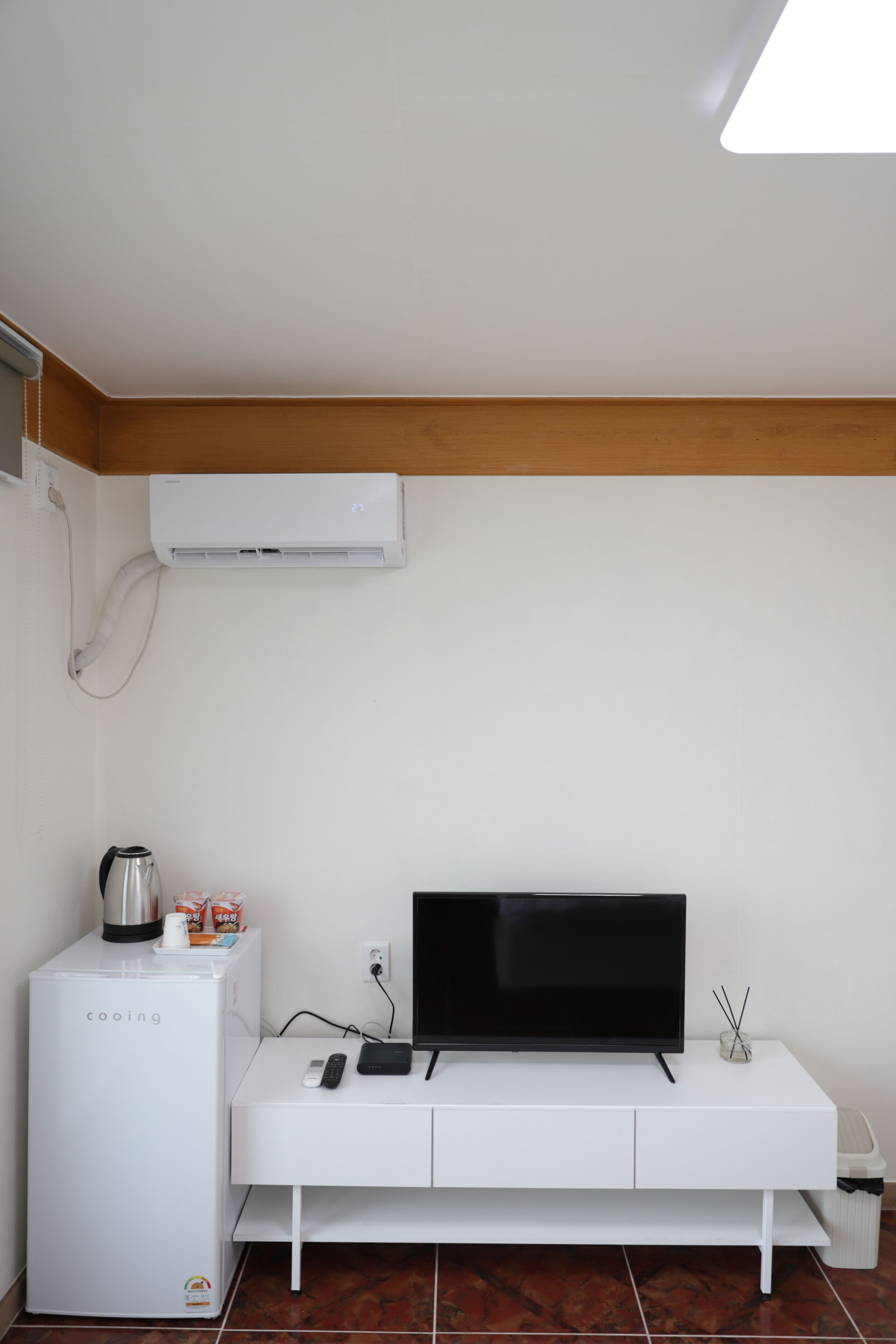 A room with a white entertainment unit, a flat-screen TV, a small refrigerator, and an air conditioning unit mounted on the wall.