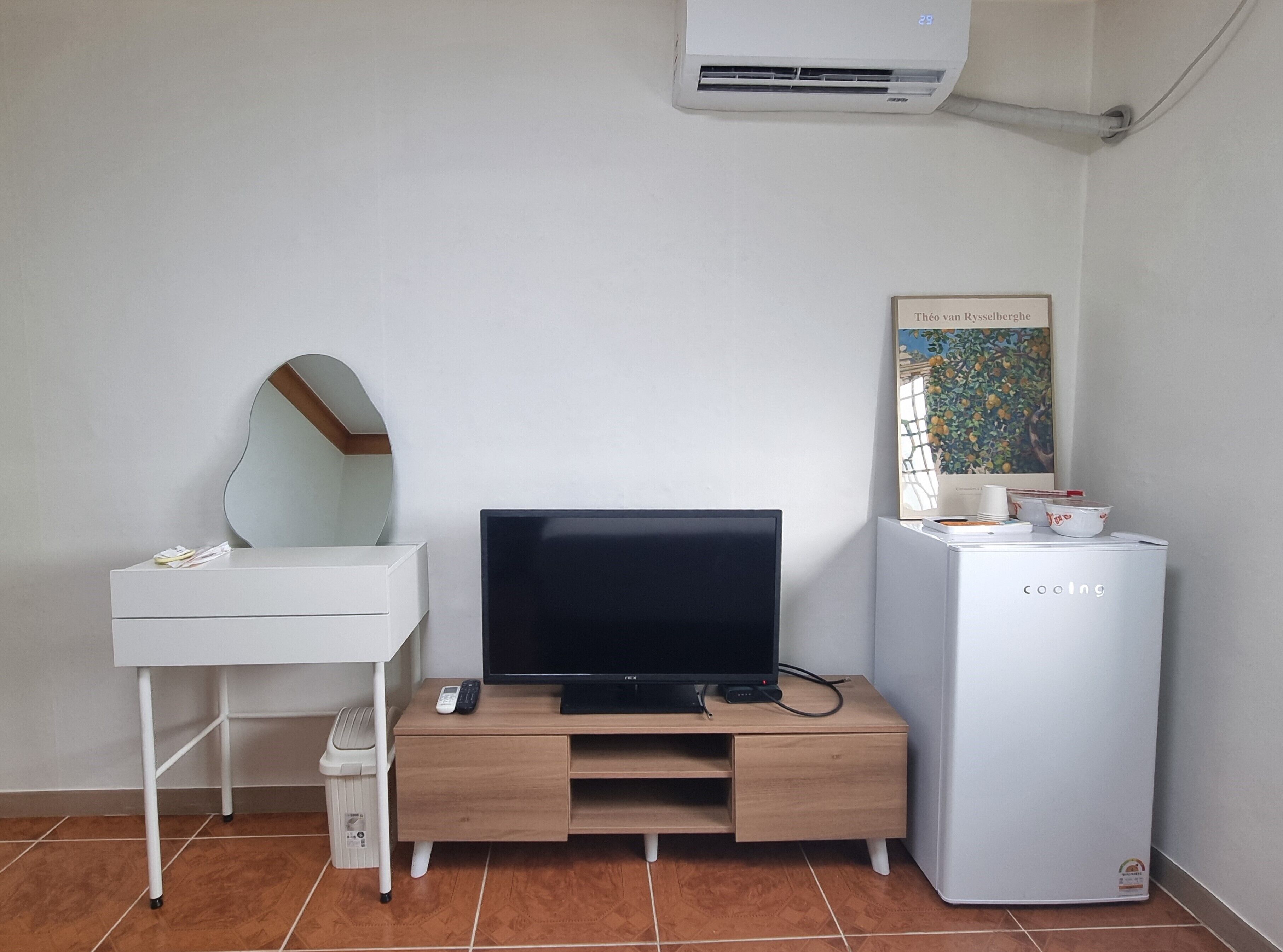 Ein Zimmer mit weißem Schreibtisch, Flachbildfernseher, kleinem Kühlschrank, Mülleimer und Wandklimaanlage.