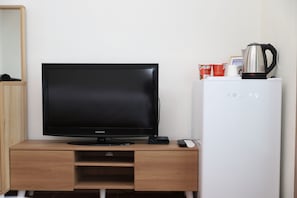 A room with a flat-screen TV on a wooden desk, a white mini-fridge with a kettle, and a remote control. - Gloriastay (Pohang)