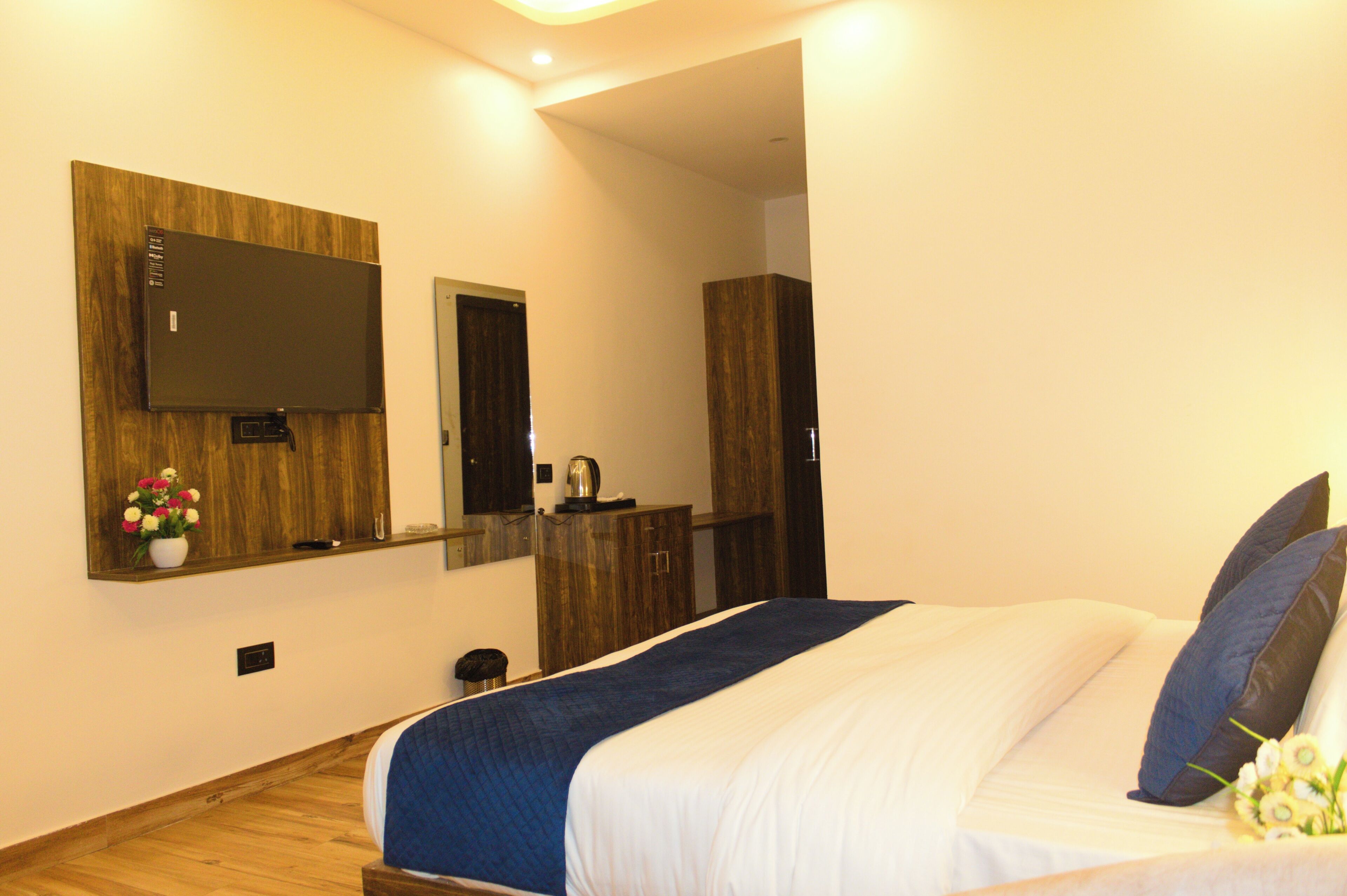 A hotel room with a bed, a flat-screen TV mounted on the wall, a wooden wardrobe, and a small table with a kettle.
