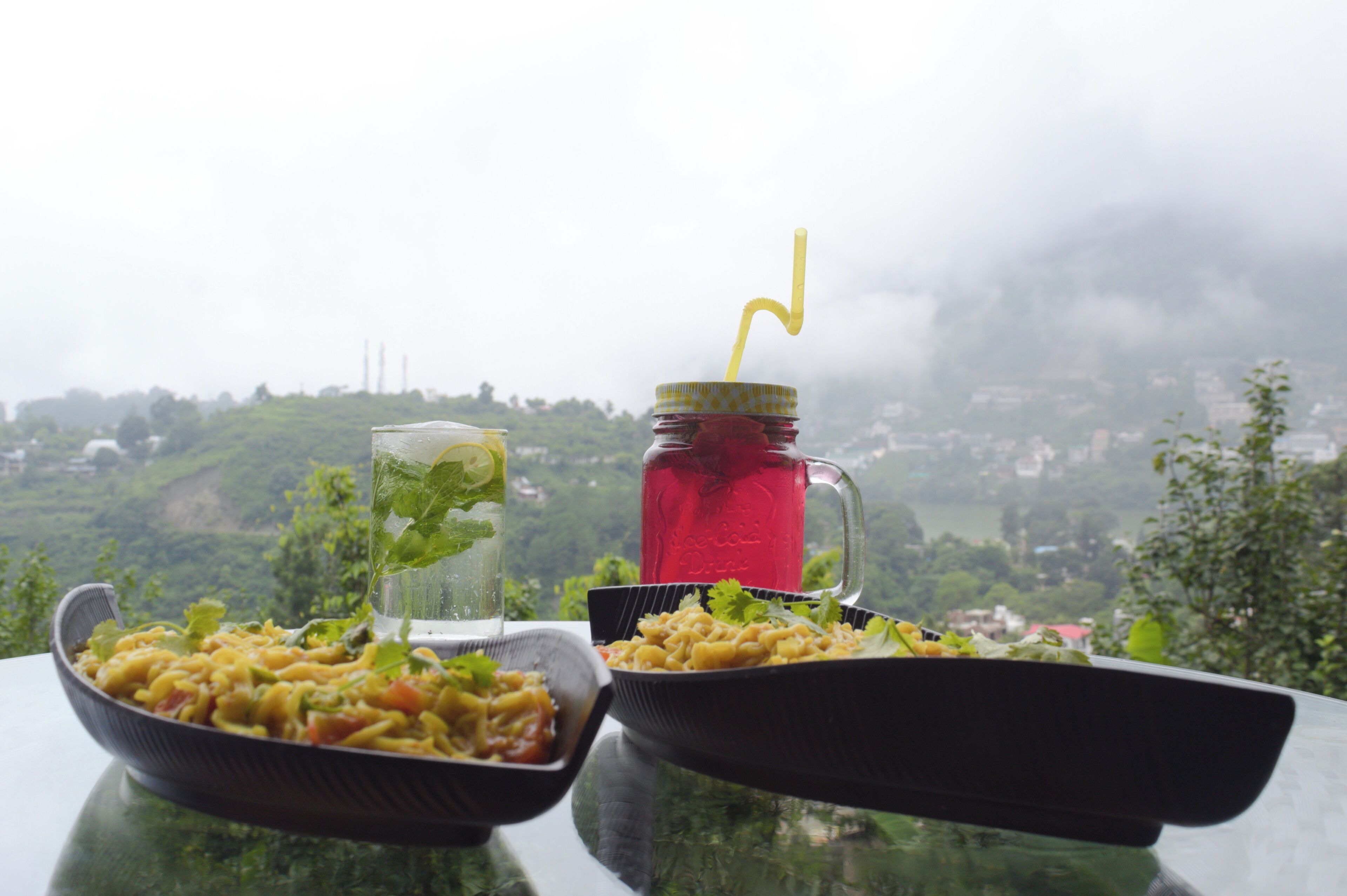 Una mesa con dos platos de pasta y un vaso de bebida helada adornado con hojas de menta, frente a una vista panorámica de un paisaje montañoso.