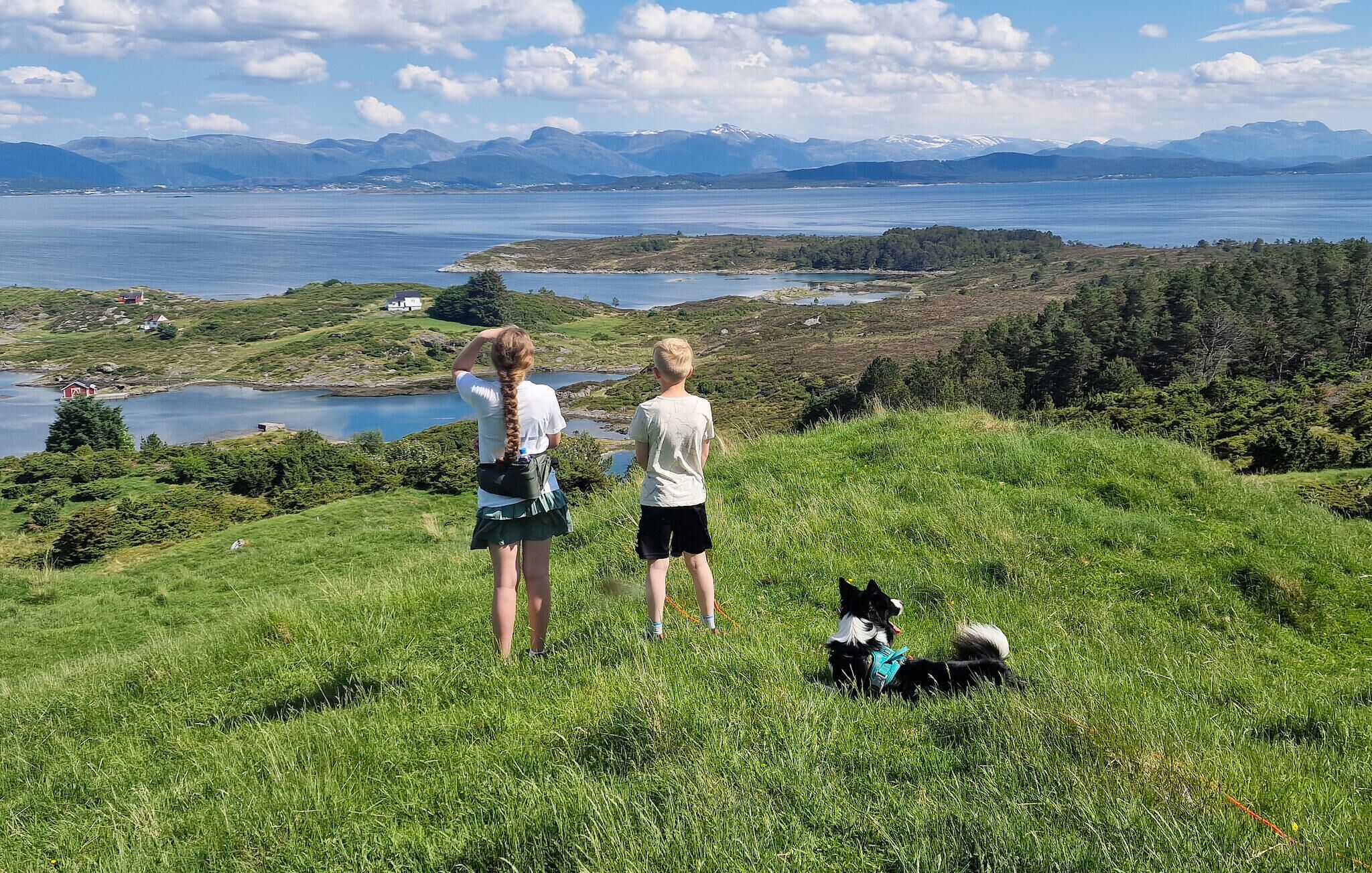 Two people and a dog on a grassy hill overlooking a body of water and mountains.