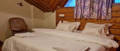 Un dormitorio con techo de madera, una cama con ropa blanca, una silla de mimbre y un ventanal con cortinas estampadas.