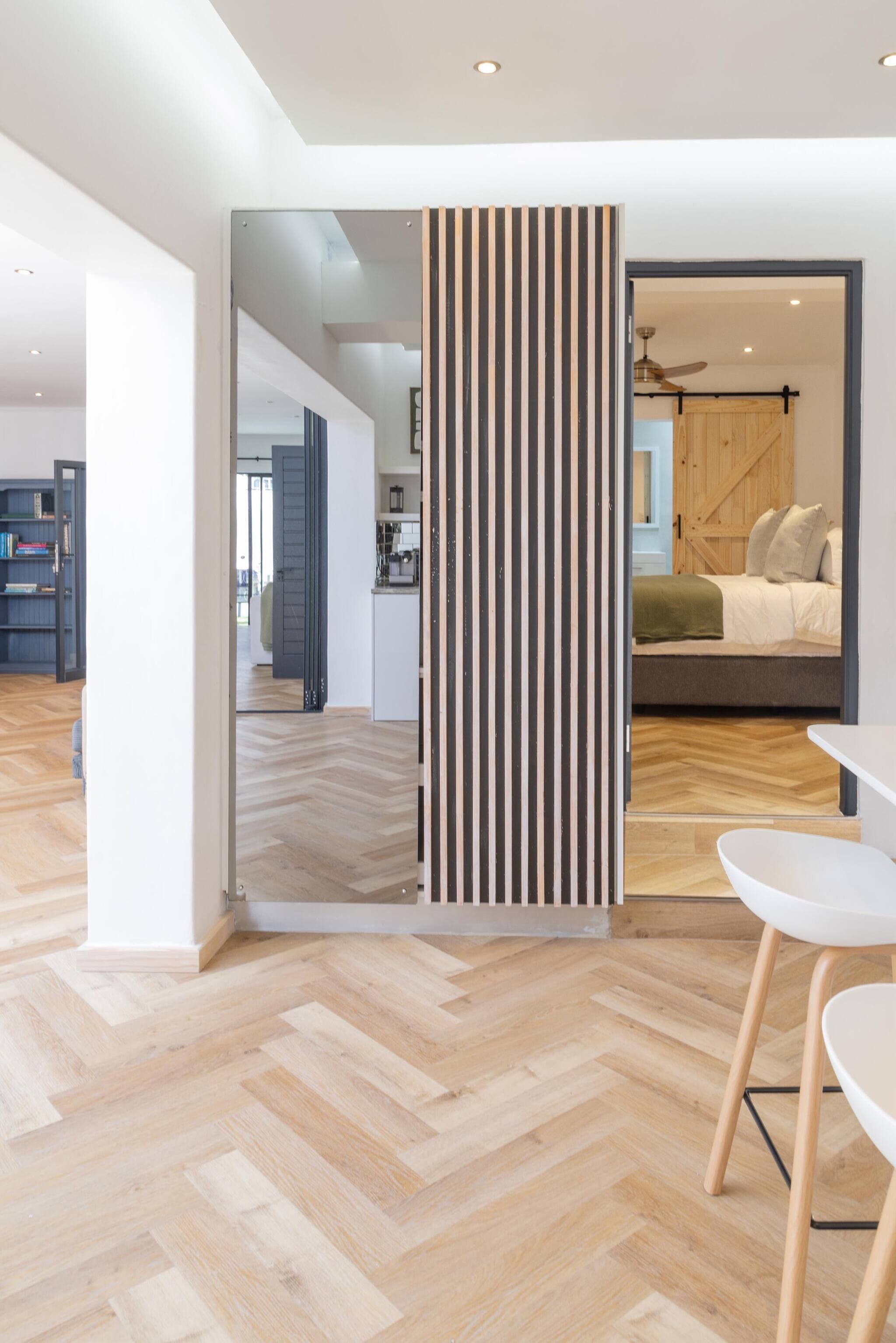 A modern interior with a herringbone wooden floor, a dining area with white chairs, and a kitchen visible in the background.