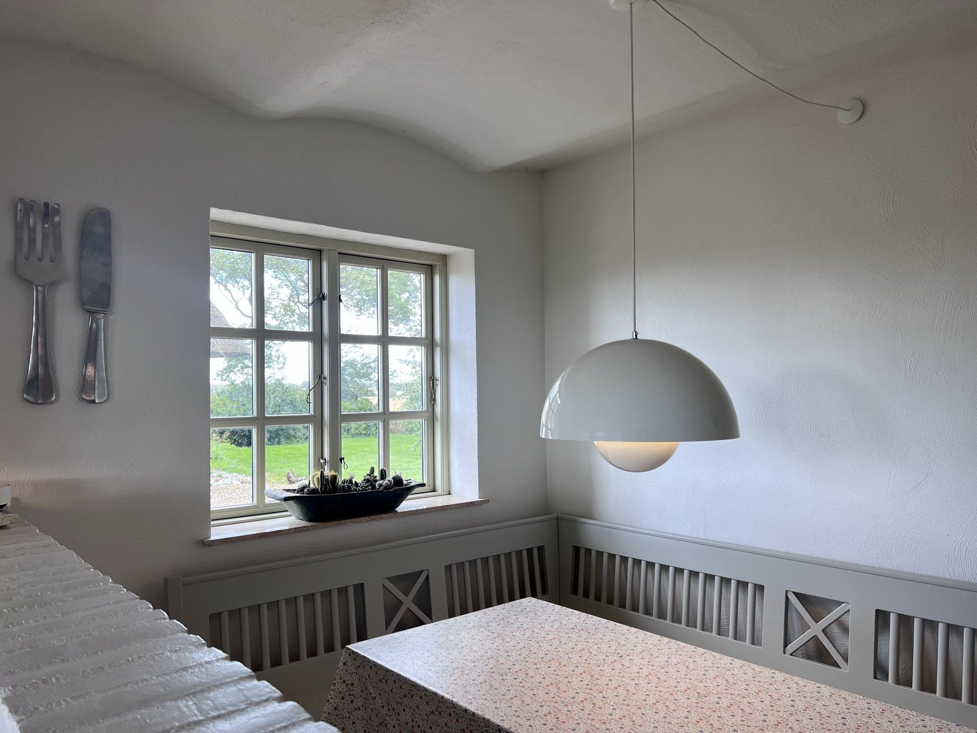 A room with a dining table, a window with a view of greenery, a hanging light fixture, and a wall-mounted knife and fork set.