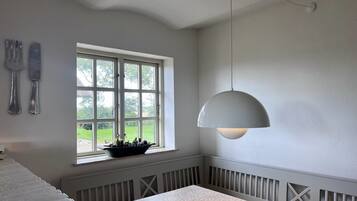 A room with a dining table, a window with a view of greenery, a hanging light fixture, and a wall-mounted knife and fork set.
