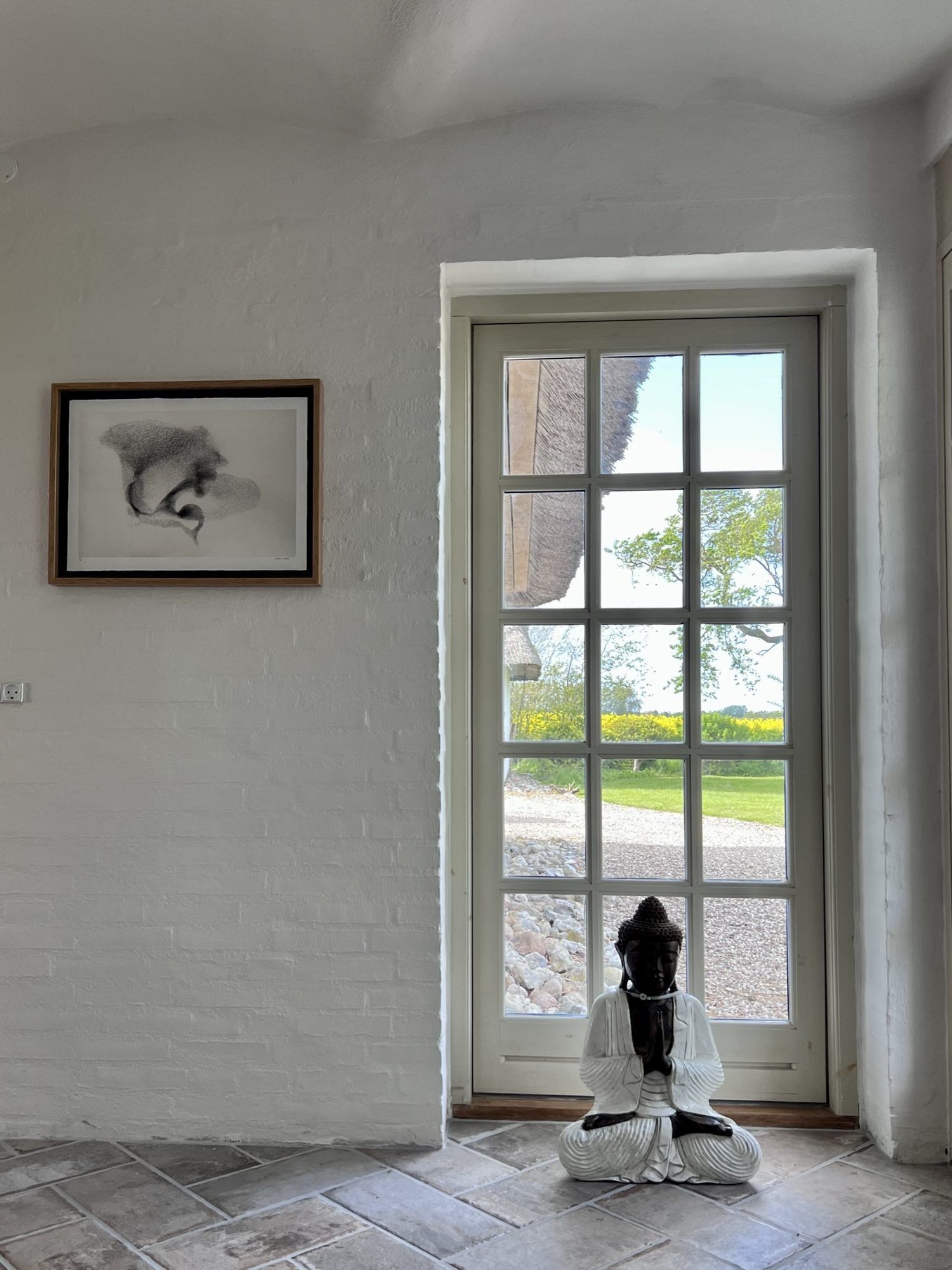 Et værelse med et stort vindue, et billede i ramme på væggen og en statue af en siddende figur.
